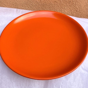 Könnte beinhalten: Ein großer, oval geformter, orangefarbener Teller mit glänzendem Finish. Der Teller steht auf einer weißen, strukturierten Oberfläche. Die leuchtende Farbe und das schlichte Design des Tellers machen ihn zu einem auffälligen Stück zum Servieren von Speisen.