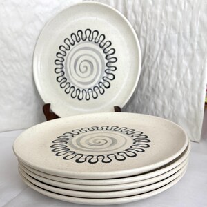 May include: A stack of five round ceramic plates with a speckled off-white finish. Each plate features a central grey spiral design surrounded by a black wavy line pattern. One plate is displayed upright on a stand behind the stacked plates.