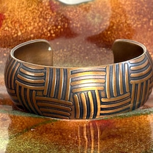 Pode incluir: Uma pulseira de punho em tom de cobre com um padrão tecido. O design aberto da pulseira apresenta linhas que se cruzam em uma cor bronze escuro, criando uma superfície texturizada. A pulseira é exibida em uma superfície reflexiva com tons quentes e terrosos.
