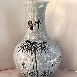 May include: A light gray ceramic vase with a prominent crackle glaze finish. The vase features a bulbous body and a flared neck. Black ink illustrations of bamboo stalks and leaves decorate the exterior, accompanied by dark brown calligraphy.