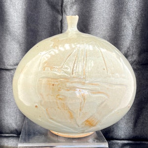 Op de afbeelding: Een ronde, gebroken witte keramische vaas met een zeilbootontwerp. De vaas heeft een bolvormig lichaam en een smalle hals. De zeilboot is in het oppervlak geëtst, met details van de zeilen en de romp. De vaas is een decoratief item.