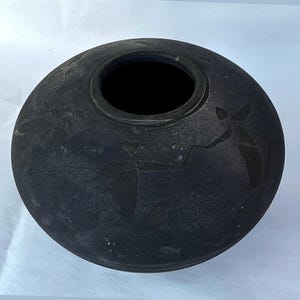 Puede incluir: Un jarrón de cerámica redondo de color gris oscuro con una abertura ancha. El jarrón tiene una superficie texturizada y diseños grabados de figuras. El jarrón mide aproximadamente 20 cm de diámetro y 15 cm de alto.