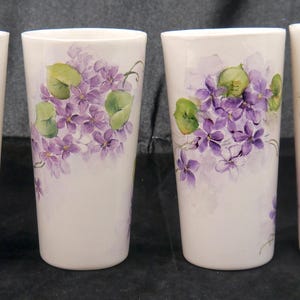 May include: A set of four white ceramic tumblers, each featuring a hand-painted floral design of purple violet flowers and light green leaves. The cylindrical vessels have a smooth finish and display varying arrangements of the botanical motif.