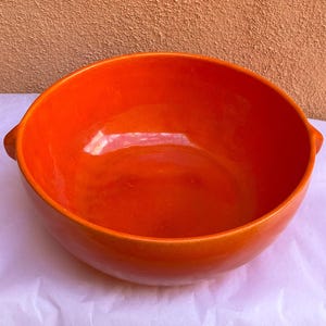 Puede incluir: Un cuenco de cerámica naranja brillante con acabado brillante. El cuenco tiene forma redondeada con dos pequeñas asas a cada lado. El interior y el exterior son del mismo color naranja brillante. Aproximadamente 25 cm de diámetro.