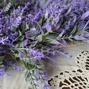May include: A close-up of a purple lavender wreath with green leaves. The wreath is lying on a white crocheted surface.