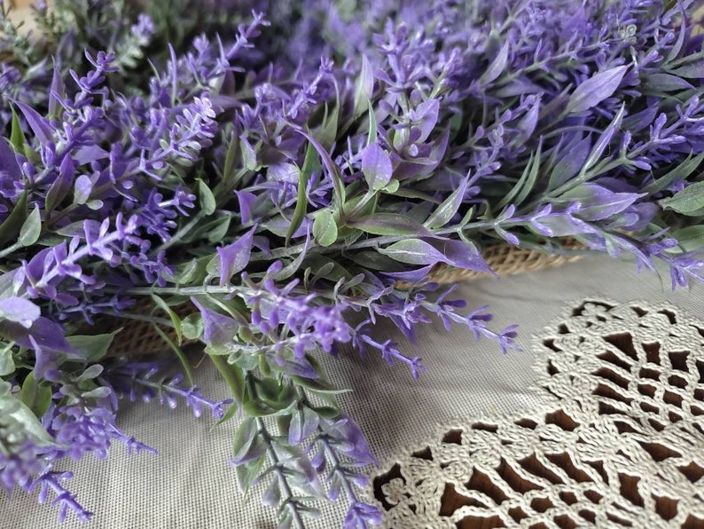 May include: A close-up of a purple lavender wreath with green leaves. The wreath is lying on a white crocheted surface.