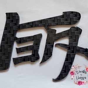 Könnte beinhalten: Schwarze, stilisierte japanische Schriftzeichen mit einem strukturierten Muster. Die Zeichen sind ausgeschnitten und scheinen aus einem dunklen Material zu bestehen. Der Hintergrund ist weiß. Das Bild enthält auch den Text "Wreaths by Vongsa" in einem floralen Design.