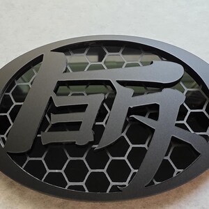 May include: An oval-shaped black emblem featuring a central Japanese kanji character layered over a dark grey honeycomb pattern. The design is framed by a solid black border, and a small colorful graphic with the text VONGSAL is in the corner.