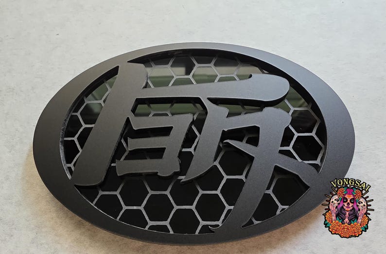May include: An oval-shaped black emblem featuring a central Japanese kanji character layered over a dark grey honeycomb pattern. The design is framed by a solid black border, and a small colorful graphic with the text VONGSAL is in the corner.