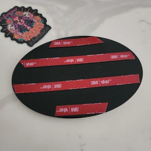 May include: A black oval-shaped plaque featuring four horizontal strips of red 3M VHB adhesive tape on its back surface. Beside the plaque is a decorative sticker depicting a stylized figure with floral elements and skulls in vibrant colors.