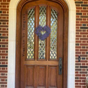 May include: A wooden door with a diamond-shaped glass window pane and a lavender heart-shaped wreath. The door has a brass door handle and is surrounded by brick and white trim. The text "Wreaths by Vongsai" is written on the door. The text "customers photo" is written at the bottom of the image.