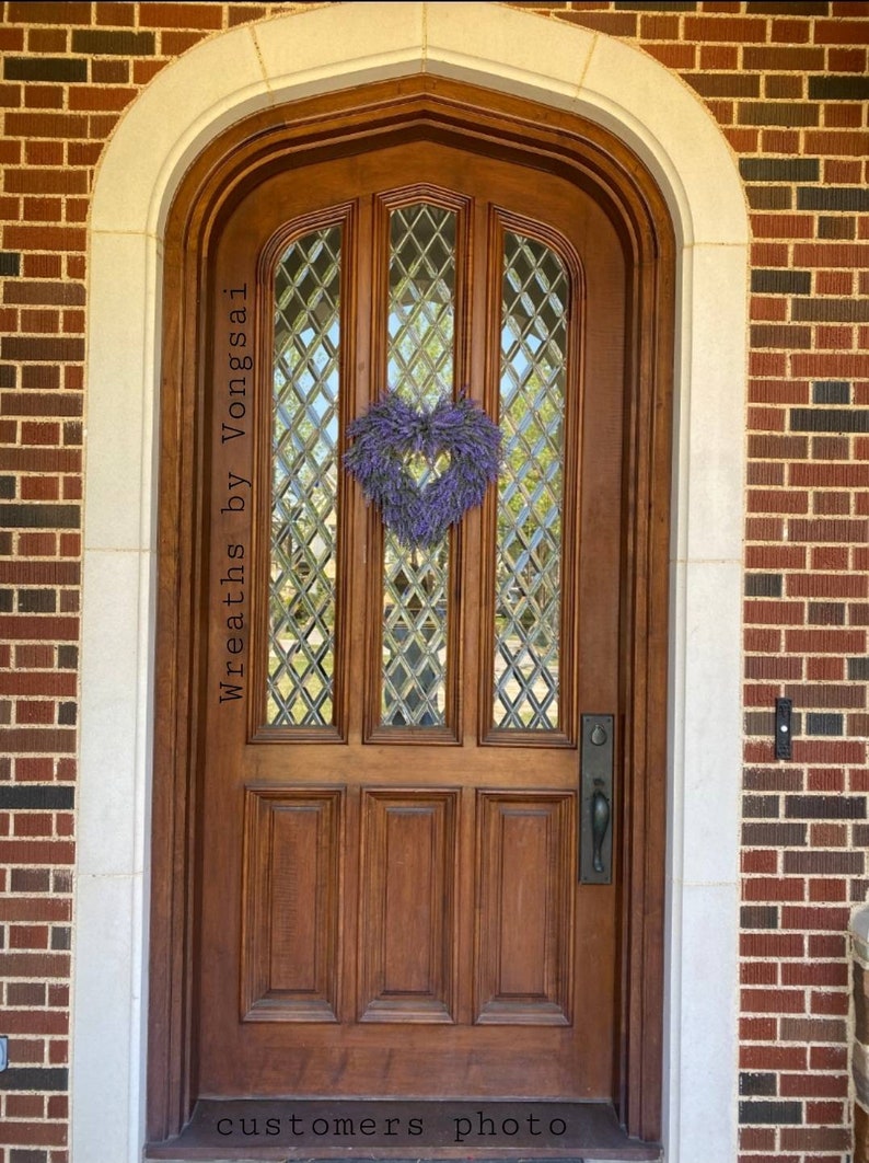 May include: A wooden door with a diamond-shaped glass window pane and a lavender heart-shaped wreath. The door has a brass door handle and is surrounded by brick and white trim. The text "Wreaths by Vongsai" is written on the door. The text "customers photo" is written at the bottom of the image.