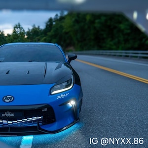 May include: A blue sports car with a black hood and front bumper, featuring white Japanese text decals on the bumper and windshield, a black grille with a glowing light strip, and bright blue underglow lighting on the asphalt road surface.