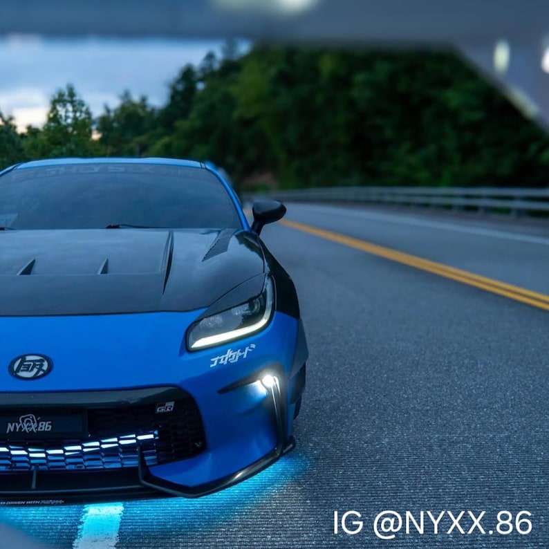 May include: A blue sports car with a black hood and front bumper, featuring white Japanese text decals on the bumper and windshield, a black grille with a glowing light strip, and bright blue underglow lighting on the asphalt road surface.