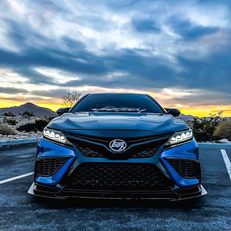 May include: A blue sedan viewed from the front, featuring a black honeycomb grille, sharp LED headlights, a custom emblem on the hood, and a white decal on the windshield. The vehicle has a low-profile front splitter and a dark, aggressive bumper design.