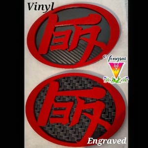May include: Two oval emblems featuring bright red Japanese kanji characters set against dark backgrounds. The top emblem has a carbon fiber pattern, while the bottom emblem displays a woven texture. The text Vinyl and Engraved are visible.