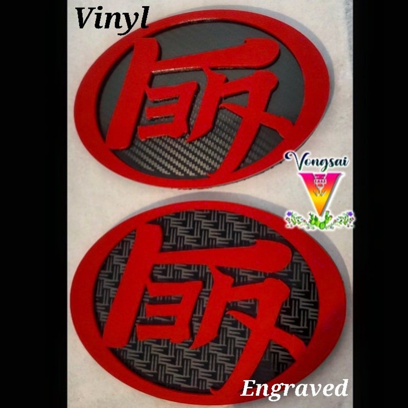 May include: Two oval emblems featuring bright red Japanese kanji characters set against dark backgrounds. The top emblem has a carbon fiber pattern, while the bottom emblem displays a woven texture. The text Vinyl and Engraved are visible.