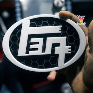Op de afbeelding: Wit ovaal embleem met de letters "FTG" in een vet, gestileerd lettertype. De letters staan tegen een zwart honingraatpatroon in het ovaal. Het embleem wordt in een hand gehouden, met een vage achtergrond.