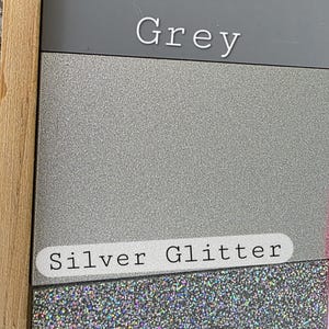May include: A rectangular display board featuring three distinct material samples. The top section is a solid matte grey, the middle is a textured silver glitter, and the bottom is a multi-colored holographic confetti glitter. Each section is labeled.