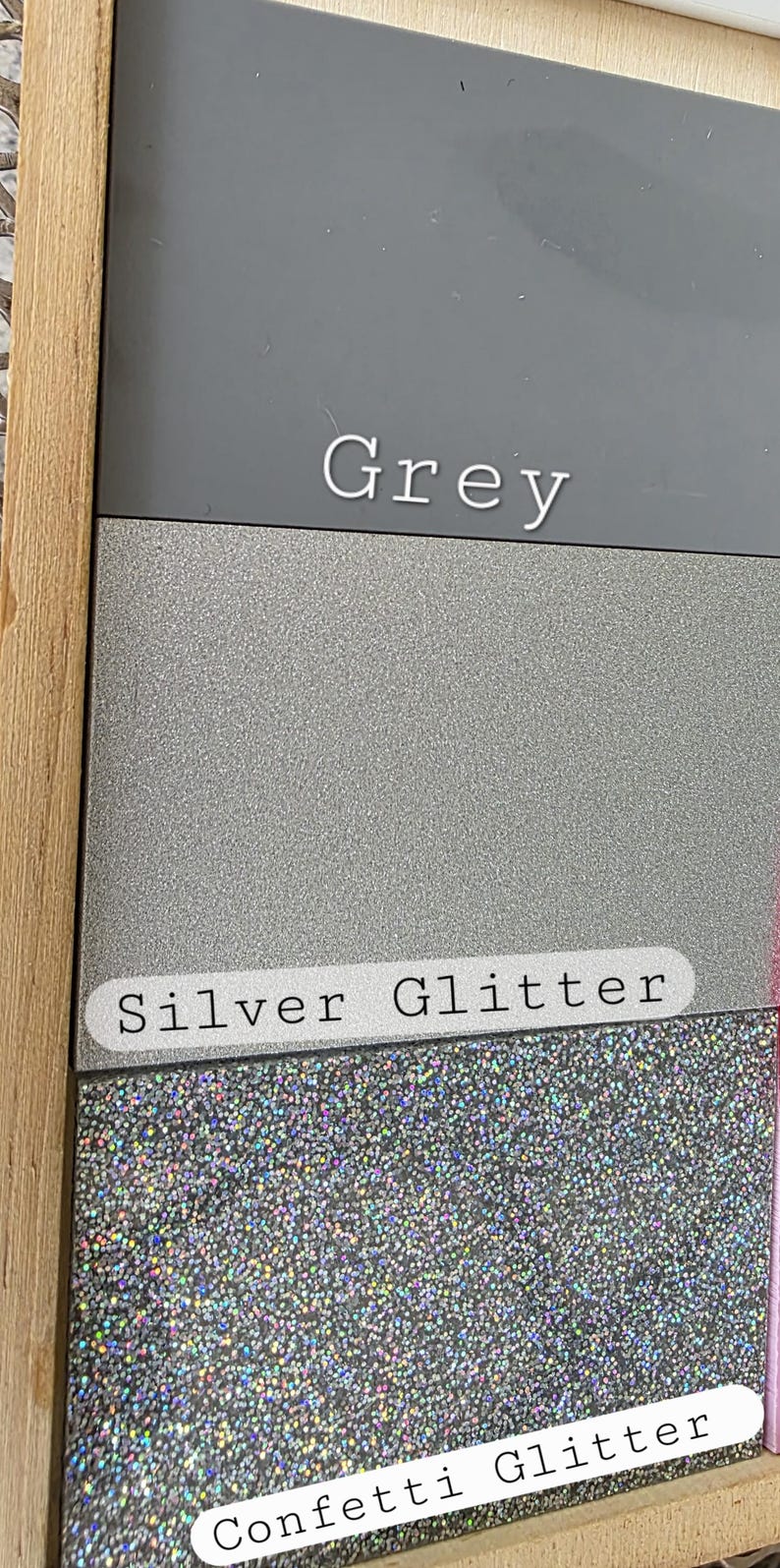 May include: A rectangular display board featuring three distinct material samples. The top section is a solid matte grey, the middle is a textured silver glitter, and the bottom is a multi-colored holographic confetti glitter. Each section is labeled.