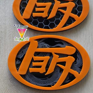 May include: Two oval-shaped orange emblems featuring raised orange Japanese kanji characters. The top emblem has a black honeycomb background, while the bottom emblem displays a dark grey dragon motif. The text Vongsai is visible on the left.