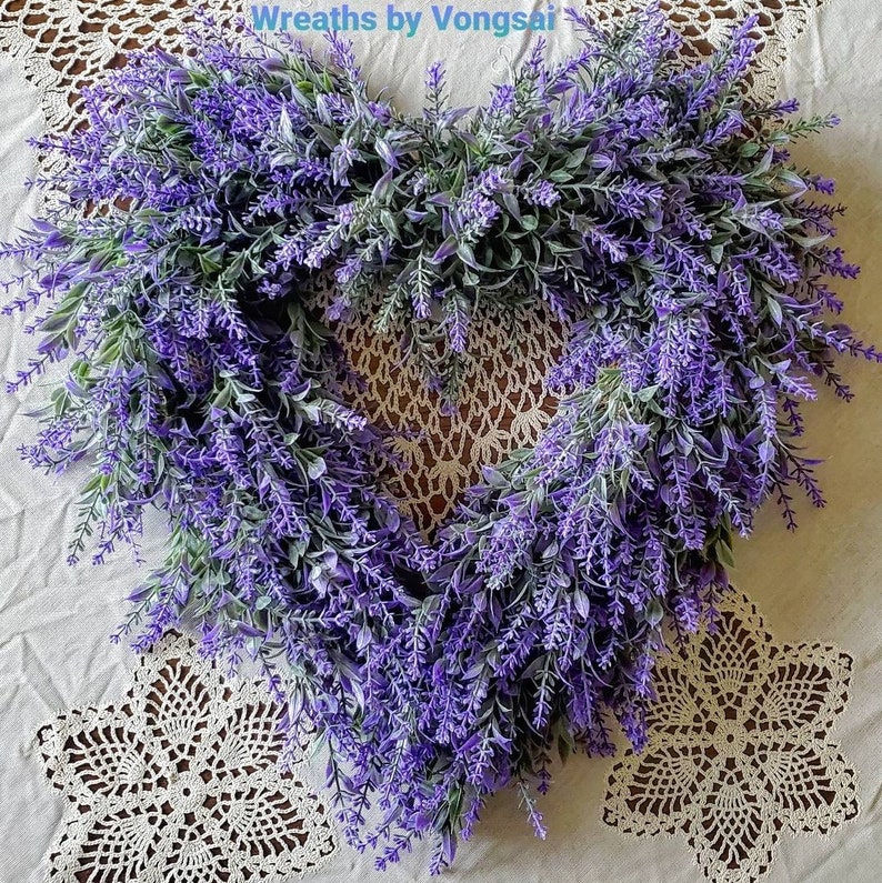 May include: A heart-shaped lavender wreath with purple flowers and green leaves. The wreath is on a white crocheted doily.