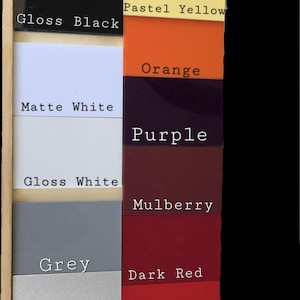 May include: A rectangular wooden display board featuring sixteen square color swatches labeled with text: Matte Black, Gloss Black, Matte White, Gloss White, Grey, Silver Glitter, Confetti Glitter, Yellow, Pastel Yellow, Orange, Purple, Mulberry, Dark Red, Red, and Pastel Pink.