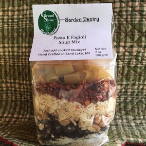 May include: A clear plastic bag filled with ingredients for Pasta E Fagioli soup mix. The bag has a label that reads "Garden Pantry" and "Pasta E Fagioli Soup Mix". The label also states "Just add cooked sausage!" and "Hand Crafted in Sand Lake, MI". The bag contains pasta, dried tomatoes, and other ingredients. The net weight is 7 oz (198 grm).