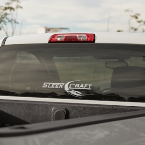 May include: White pickup truck with a black bed and a rear window sticker that says "SLEEK CRAFT" with a stylized logo of a boat.