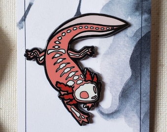 Op de afbeelding: Een roze en witte axolotl-emaille pin met zwarte contouren. De pin is op een witte kaart met zwarte inktvlekken. De kaart heeft ook de tekst "Etsy" en "@KATTAKIND" erop gedrukt.