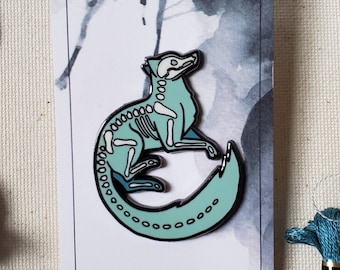Op de afbeelding: Een tealblauwe emaille pin van een skeletvos die in een cirkel is opgerold. De pin is op een witte kaart met de tekst "Etsy @KATTAKIND".
