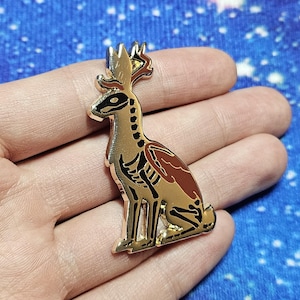 May include: A gold-coloured enamel pin depicting a stylized jackalope with a skeleton design. The jackalope has brown and black details, with a detailed antler design. The pin is set against a blue background.