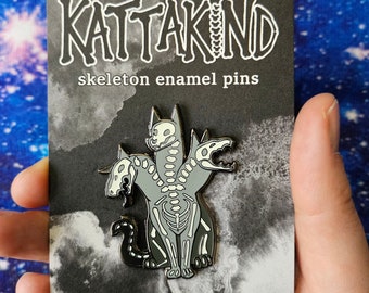 Pode incluir: Um broche de esmalte preto e branco com um gato esquelético de três cabeças sobre um fundo cinza. O broche está em um cartão com o texto "KATTAKIND skeleton enamel pins".