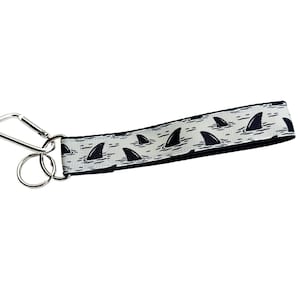 May include: A white lanyard with a repeating pattern of black shark fins emerging from the water. The lanyard has a silver metal clip and a split ring for attaching keys or an ID badge.