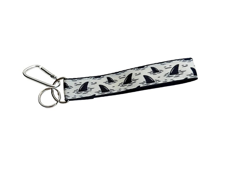 May include: A white lanyard with a repeating pattern of black shark fins emerging from the water. The lanyard has a silver metal clip and a split ring for attaching keys or an ID badge.