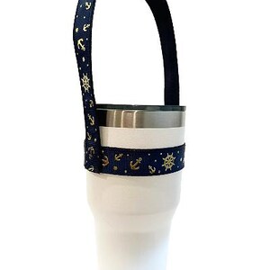 Op de afbeelding: Een witte tumbler met een donkerblauwe en gouden anker- en scheepswiel-patroonband. De band is bevestigd aan de tumbler en vormt een handvat.