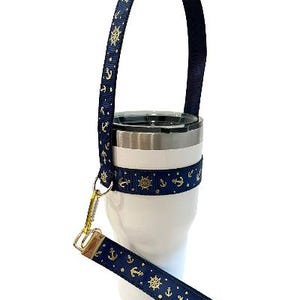 Op de afbeelding: Een witte tumbler met een blauw en gouden anker- en scheepswielontwerp. De tumbler heeft een blauwe band met een gouden sluiting.