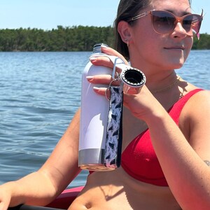 May include: A white stainless steel water bottle with a silver base and a black and white shark fin wrist strap. The bottle is held by a person wearing a red top and sunglasses, with a body of water in the background.