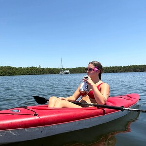 May include: A red kayak on a calm body of water with a person holding a beverage. The kayak has a black paddle and is surrounded by water and trees. The sky is blue and the sun is shining.