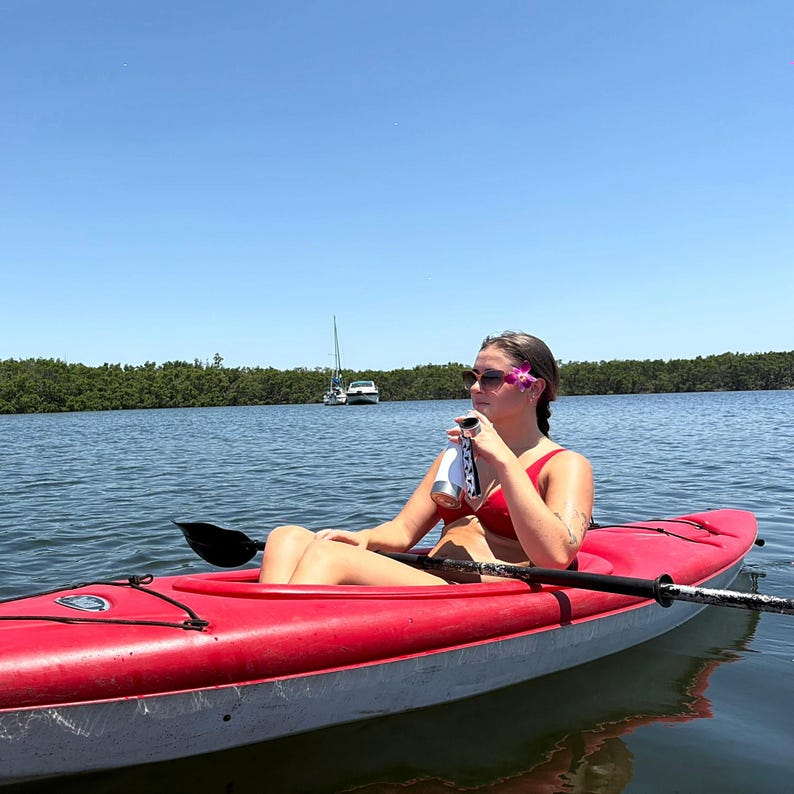 May include: A red kayak on a calm body of water with a person holding a beverage. The kayak has a black paddle and is surrounded by water and trees. The sky is blue and the sun is shining.