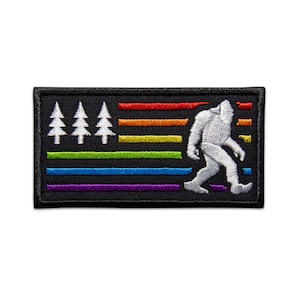 May include: A black embroidered patch featuring a white silhouette of Bigfoot walking across a rainbow-coloured flag with white pine trees on the left side.