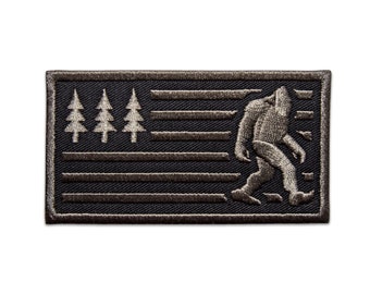 May include: A black and grey embroidered patch featuring a Bigfoot walking across a stylised American flag with three pine trees in the top left corner.