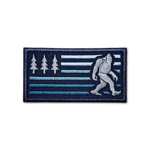 May include: A blue and white embroidered patch with a stylized image of Bigfoot walking across a flag with a row of pine trees on the left side.