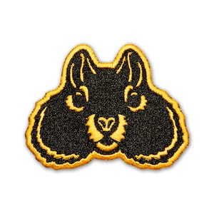 Könnte beinhalten: Ein gestickter Aufnäher in Schwarz und Gelb, der das Gesicht eines Eichhörnchens zeigt. Das Eichhörnchen hat große Ohren, eine kleine Nase und einen buschigen Schwanz.
