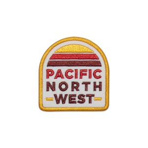 Könnte beinhalten: Ein weißer gestickter Aufnäher mit gelbem Rand. Der Aufnäher zeigt einen stilisierten Sonnenuntergang mit roten, orangen und braunen Streifen. Der Text "PACIFIC NORTH WEST" ist in braunen Buchstaben unter dem Sonnenuntergang geschrieben.