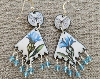 May include: A pair of dangle earrings with a floral theme. Each earring features a white, triangular enamel pendant with a blue flower and green leaves. A small, round pendant with a butterfly tops each earring. Blue crystal beads dangle from the bottom.