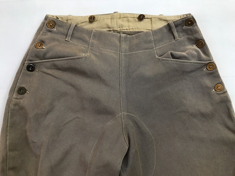 May include: A pair of brown twill pants with a button fly and multiple button closures along the sides. The pants have a high waist and a straight leg.