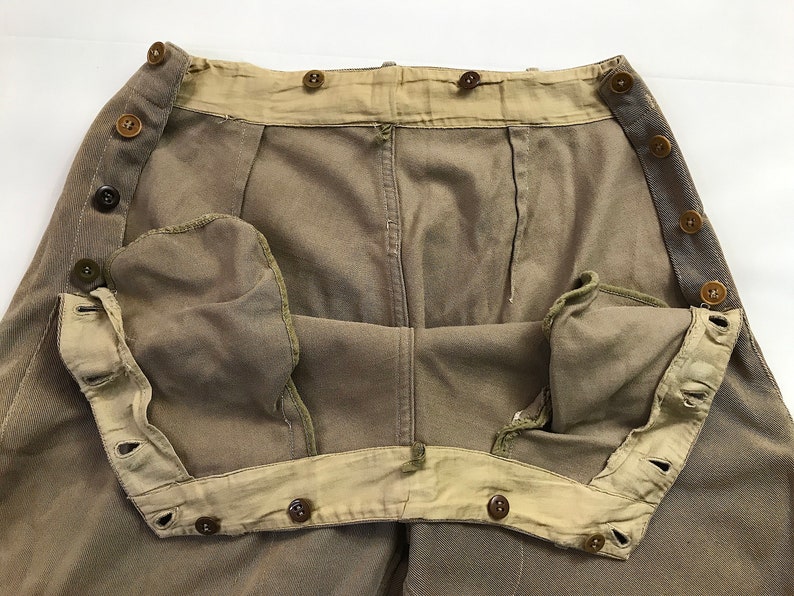 May include: A pair of brown twill pants with a button fly and a buttoned waistband. The pants have a light brown lining and are turned inside out to show the lining.
