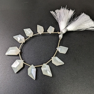 Op de afbeelding: Een streng witte maansteen kralen met een facetgeslepen, diamantvormig ontwerp. De kralen zijn geregen op een zilveren draad en hebben een witte kwast aan het einde.