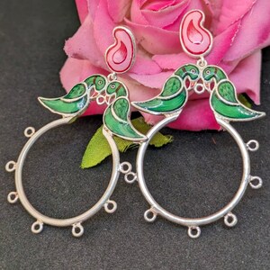 May include: A pair of silver hoop earrings with green and pink enamel detailing. The earrings feature a design of two birds facing each other, with a pink paisley shape hanging from the top of each hoop.
